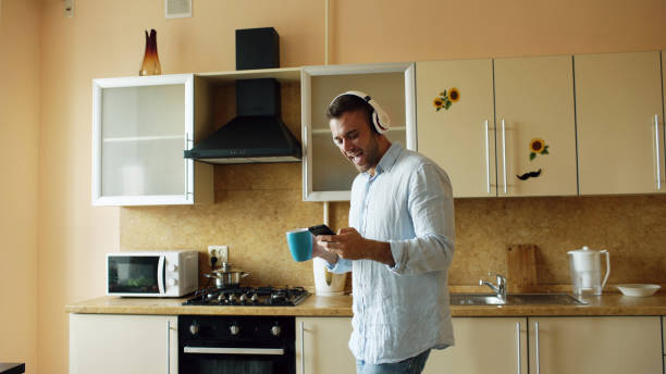 Handsome young funny man in headphones dancing and singing in kitchen at home in the morning and have fun on vacation stock photo