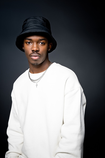 Fashion portrait of young man wearing white sweatshirt, black bucket hat, looking at camera. Studio shot on black background.