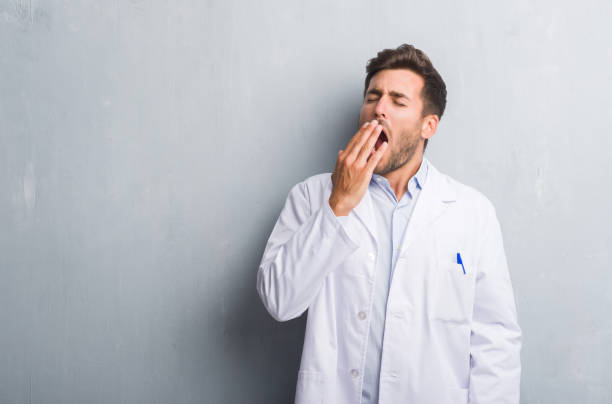 handsome young professional man over grey grunge wall wearing white coat bored yawning tired covering mouth with hand. restless and sleepiness. - doctor wall imagens e fotografias de stock