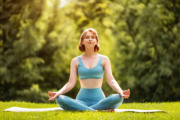 Happy adorable sporty girl performing yoga while sitting on mat in lotus position in nature stock photo