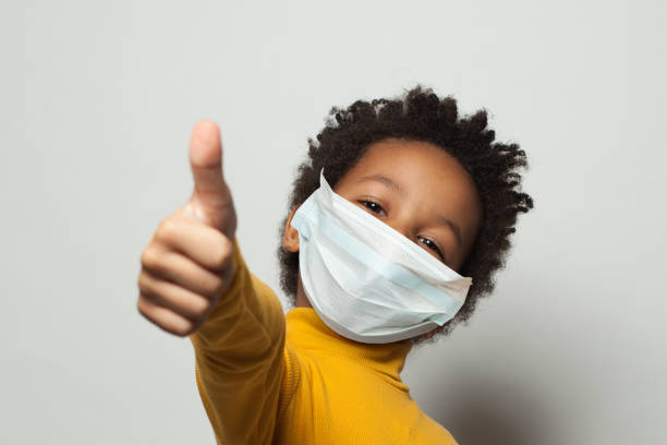 Happy African American black kid in medical protective face mask showing thumb up on white  pandemic illness stock pictures, royalty-free photos & images