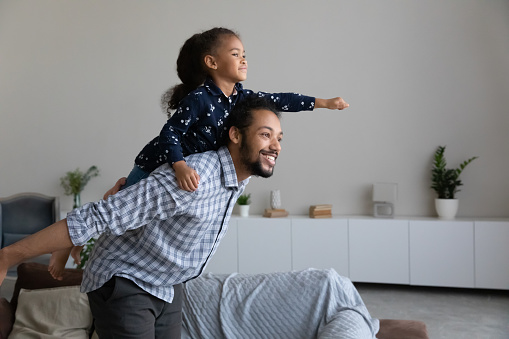 Happy African American dad piggybacking excited proud daughter kid, playing flying superhero, reaching arm forward. Cheerful girl riding fathers back playing active game with daddy at home