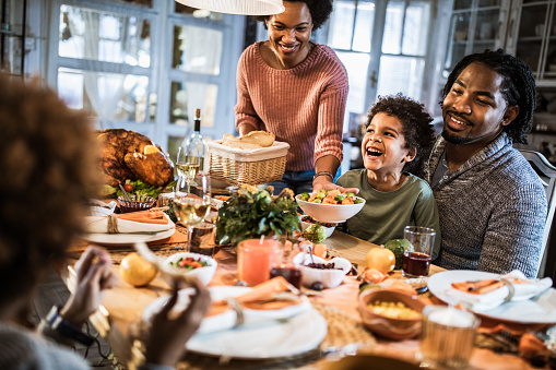 Happy black mother serving dish to her family during Thanksgiving meal at dining table.
