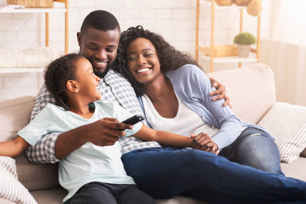 Happy african american family relaxing and watching TV at home Smiling african american family relaxing on couch and watching TV at home, man switching channels comfortable stock pictures, royalty-free photos & images