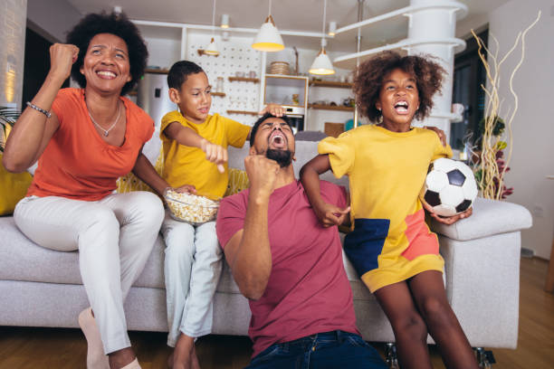Happy african american family watching soccer match on television in living room at home. stock photo