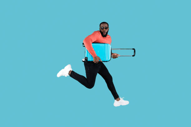 Happy African American man in a sunglasses and in casual wear jumping with a suitcase in his hands on a blue background. In anticipation of travel stock photo