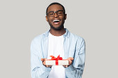 Happy african american male wearing glasses isolated on grey studio background hold wrapped birthday present with red bow, smiling black man in spectacles greeting congratulating give gift box