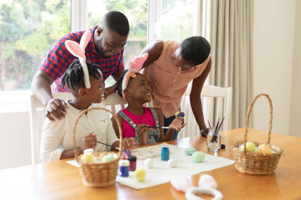 Happy african american parents with son and daughter wearing bunny ears painting colourful eggs stock photo