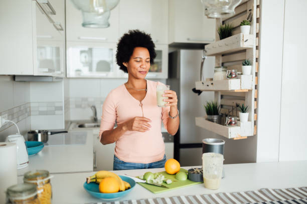 Happy African American woman preparing a nutritional breakfast at home Cheerful African American woman having a healthy breakfast at home, making a shake out of different kinds of fruits and smiling drinking smoothie stock pictures, royalty-free photos & images