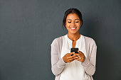 Mid adult african american woman texting message on smart phone isolated on grey background. Smiling indian woman using mobile phone and looking at it. Happy casual multiethnic lady messaging on the smartphone with new app.