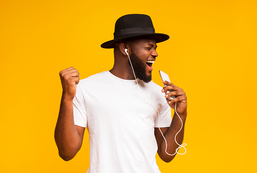 Emotional african guy in black hat singing into smartphone like microphone and listening to music via earphones, yellow background with free space