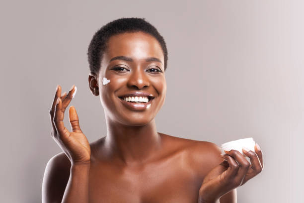 Happy Afro Woman Holding and Applying Moisturizing Cream on Face Skin Hydration. Portrait Of Cheerful Black Girl Holding and Applying Moisturizing Cream on Face, Grey Background With Free Space human skin stock pictures, royalty-free photos & images