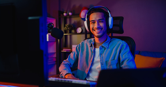Happy asia guy gamer wear headphone participation play video game colorful neon lights computer and smiling look at camera in living room at night modern house. Esport streaming game online.