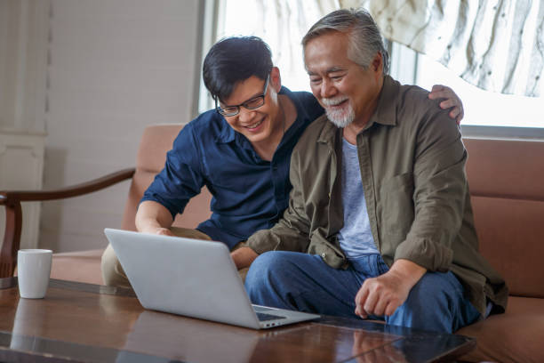 happy asian adult son and senior father sitting on sofa using laptop together at home . young man teaching old dad using internet online with computer on couch in living room . stock photo