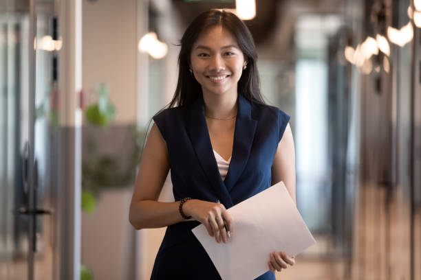 Happy asian businesswoman looking at camera stand in office hallway Happy smiling asian businesswoman looking at camera holding papers stand in office hallway, happy confident chinese professional executive satisfied with good career posing in modern office, portrait human resources photos stock pictures, royalty-free photos & images