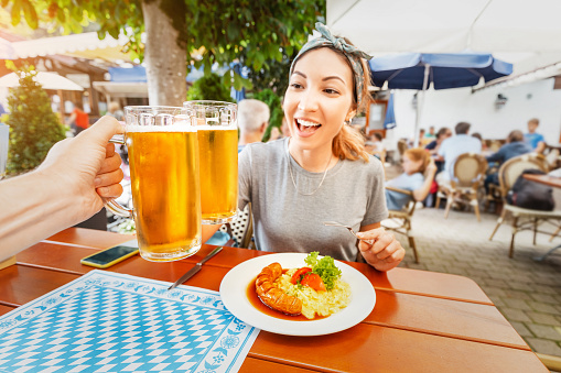 Happy Asian girl clinks a mug of Bavarian beer in Biergarten. Traditional German snack with sausage and potato salad