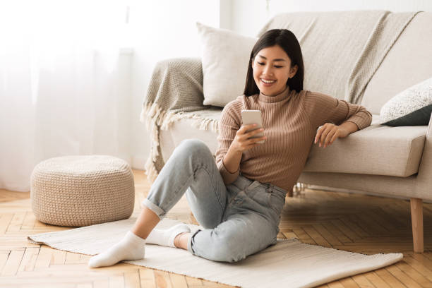 Happy Asian Girl Messaging on Phone at Home Happy Asian Girl Messaging on Phone at Home, Sitting on Floor near Sofa asian woman using phone stock pictures, royalty-free photos & images