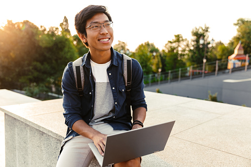 Happy asian man student with backpack using laptop computer while sitting outdoors