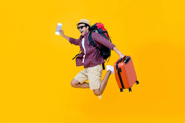 Happy Asian man with air ticket jumping Attractive happy young Asian man tourist with air ticket and passport in his hand jumping isolated on yellow studio background tourist stock pictures, royalty-free photos & images
