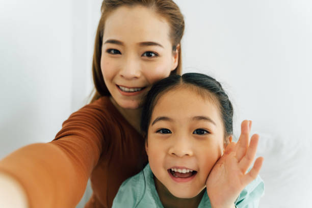 カメラに向かって手を振るかわいい女の子と幸せなアジアの母親 - 自撮り ストックフォトと画像