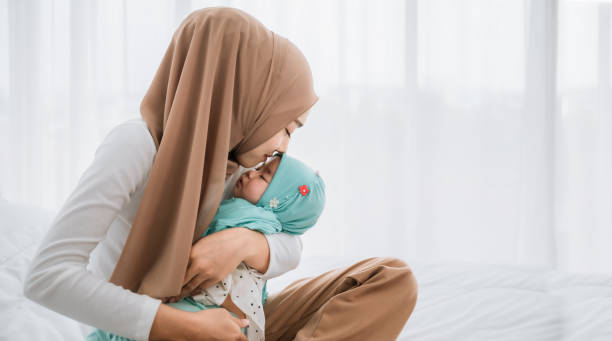 Happy asian muslim mother holding and kissing adorable little baby daughter wearing hijab in her arms on white bed in bedroom. stock photo