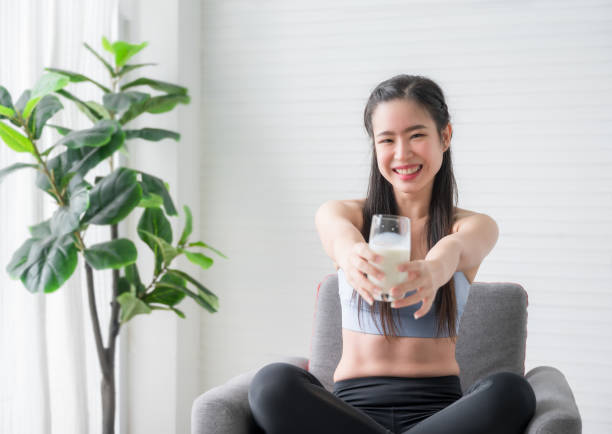 Happy Asian sports girl in sportswear sitting on chair and drinking pure milk from glass after exercise in home. stock photo