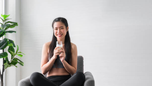 Happy Asian sports girl in sportswear sitting on chair and holding pure milk from glass after exercise in home. stock photo