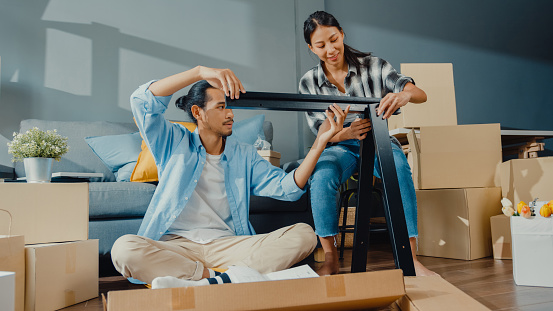 Happy asian young attractive couple man and woman help each other unpacking box and assemble furniture decorate house build table with carton box in living room. Young married asian move home concept.