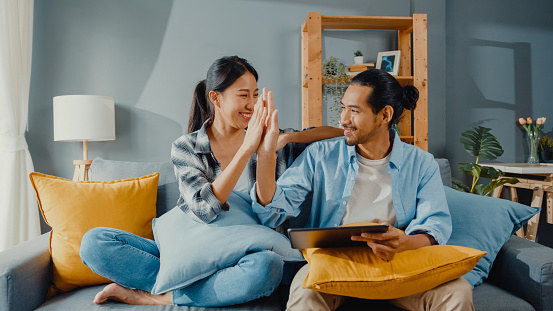 Happy asian young attractive couple man and woman sit on couch use tablet shopping online furniture decorate home in the living room at new house. Young married moving home shopper online concept.