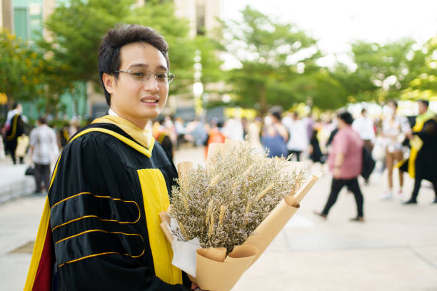Happy Asian young man in graduation ceremony. Happy Asian graduate student holding a beautiful bouquet of flowers in the university graduation ceremony. Master degree student in gown suit showing a bouquet of flower for photography. bachelor degrees stock pictures, royalty-free photos & images