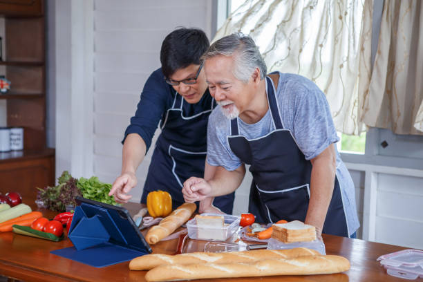 happy asian young son and senior old father cooking online class on laptop computer together making fresh vegetables food in kitchen at home stock photo