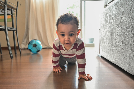 Portrait of a cute and playful baby boy crawling on the floor in the living room, looking at the camera with a beautiful smile. Behind him there is a ball, and a window open with light coming in.