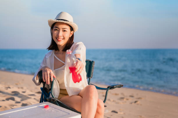 Happy beautiful Asian woman sitting on picnic chair wear sunglasses and hat holding red beverage bottle on beach in outdoor vacation summer. stock photo