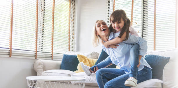 Happy Beautiful Mother carrying or piggyback her little daughter laughing playing and having fun together on sofa stock photo