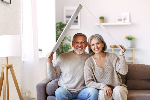Happy beautiful senior caucasian family couple holding white roof above head symbol of new home stock photo
