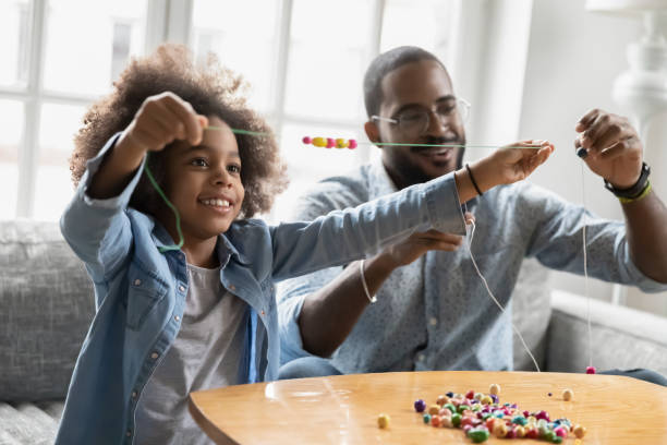 Happy biracial kid girl involved in handmade activity with father. Happy small african american kid girl involved in handmade activity with affectionate smiling biracial father at home, enjoying making wooden bracelets accessories at home, sitting at coffee table. craft stock pictures, royalty-free photos & images