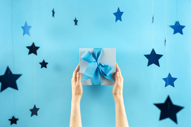 happy birthday party! photo of one big wrapped gift box with ribbon in girls hands isolated against shine and light blue background with paper stars decorations - happy birthday celebrity imagens e fotografias de stock