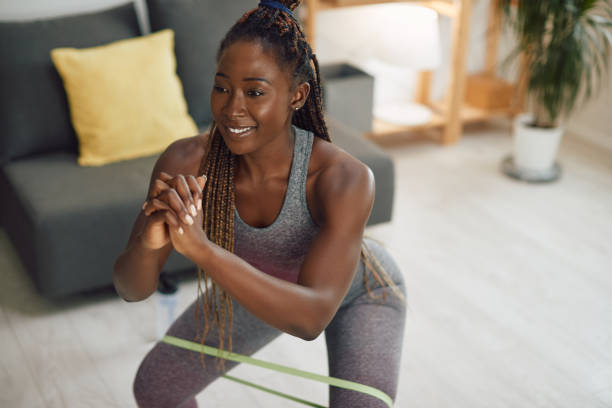 happy black athletic woman doing squats with resistance band on her around her legs at home. - träningsgummiband bildbanksfoton och bilder