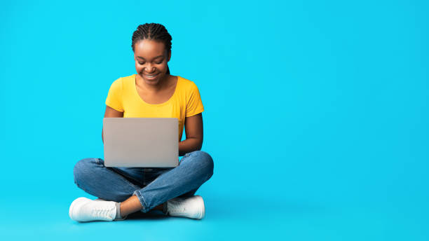 Happy Black Girl Using Laptop Sitting Over Blue Background, Panorama Happy Black Millennial Girl Using Laptop Sitting Over Blue Studio Background. Distant Learning Concept. Panorama, Copy Space sitting stock pictures, royalty-free photos & images