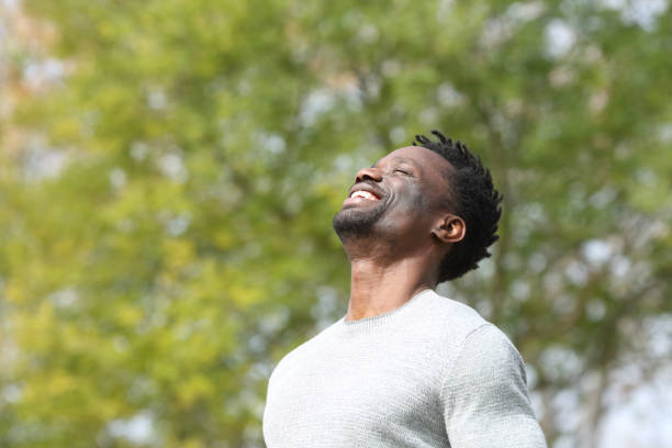 Happy black man breathing deeply fresh air in a park Happy black man breathing deeply fresh air in a park with a green tree in the background a sunny day relief emotion stock pictures, royalty-free photos & images