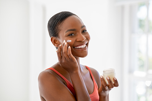 Middle aged woman applying hydrating moisturizer on her face, daily routine skin care. Smiling middle aged african woman holding little jar of skin cream and applying lotion on cheek. Beautiful young black lady applying anti-aging cream on face.