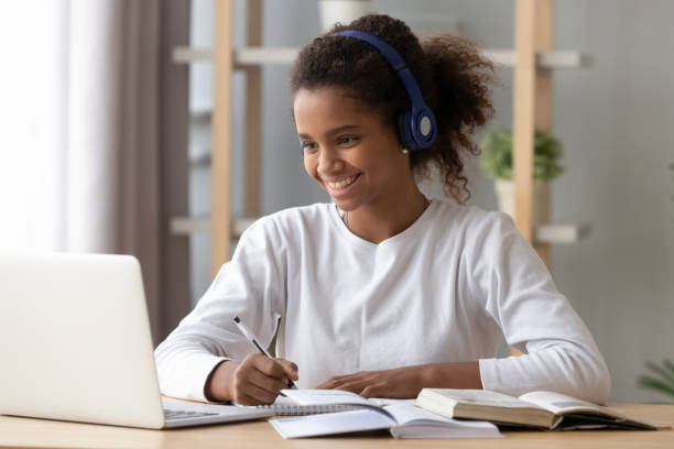 Happy black pupil in headphones doing school homework Smiling black teenage girl wearing headphones using laptop preparing homework, happy African American pupil listen to music, doing school task at home, search information online at computer homework stock pictures, royalty-free photos & images