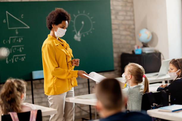 Happy black teacher giving exam paper to her student while wearing protective face mask in the classroom. Happy black teacher and her students wearing protective face mask in the classroom. Teacher is giving them their test results. lecture hall stock pictures, royalty-free photos & images