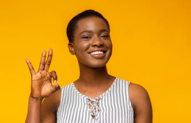 Happy black woman gesturing OK sign and smiling Everything is OK. Happy african american woman gesturing OK sign and smiling, yellow studio background, empty space sign language stock pictures, royalty-free photos & images