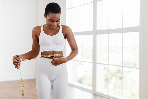 Weight Loss Concept. Portrait of smiling fit African American lady measuring her waist looking at yellow tape, blurred background. Slim black woman happy with her body. Healthy lifestyle, free space