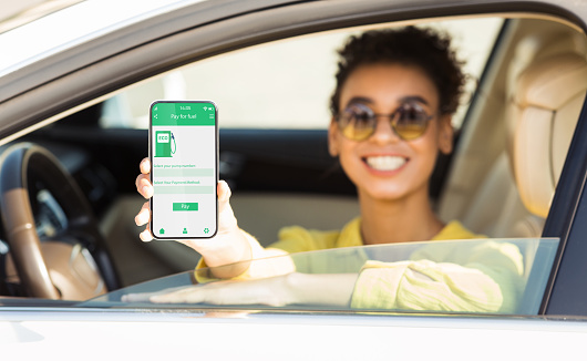 Happy black woman showing smartphone with mobile app to purchase eco fuel from her car window, panorama. African American lady choosing ecological petrol, making online payment, collage