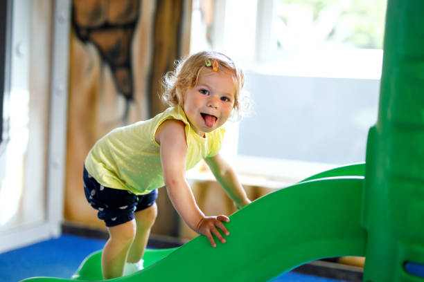 Happy blond little toddler girl having fun and sliding on indoor playground at daycare or nursery. Happy blond little toddler girl having fun and sliding on indoor playground at daycare or nursery. Positive funny baby child smiling. Healthy girl climbing on slide indoor playground stock pictures, royalty-free photos & images