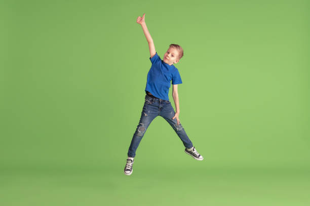 Happy boy playing and having fun on green studio background, emotions Thumb up. Happy boy playing and having fun on green studio background. Caucasian kid in bright cloth looks playful, laughting, smiling. Concept of education, childhood, emotions, facial expression. boy jumping stock pictures, royalty-free photos & images