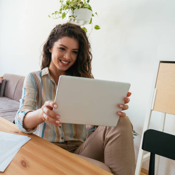 Happy Business Woman Using Digital Tablet at Home Office Cheerful smiling businesswoman talking on a video call meeting on her tablet while sitting at table with papers and documents in the living room. video call photos stock pictures, royalty-free photos & images