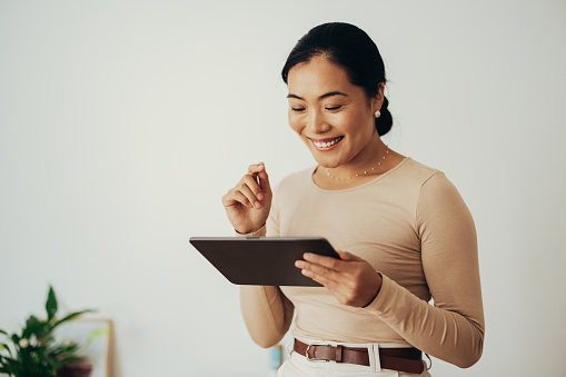 Beautiful smiling Asian businesswoman reading something on a digital tablet while standing in a modern office